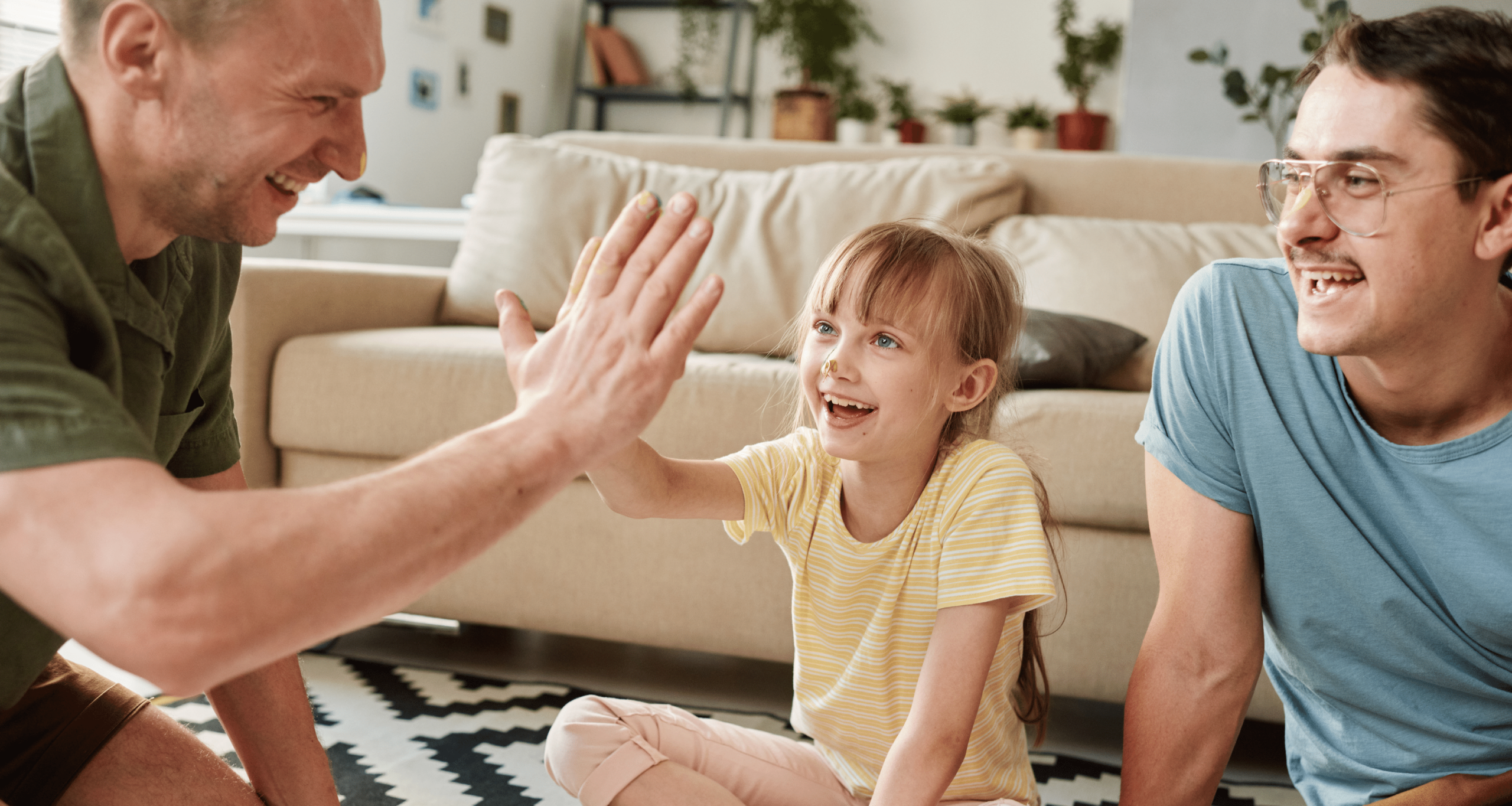 Two male adults sit on floor next to young girl while one of the adults high fives the girl 