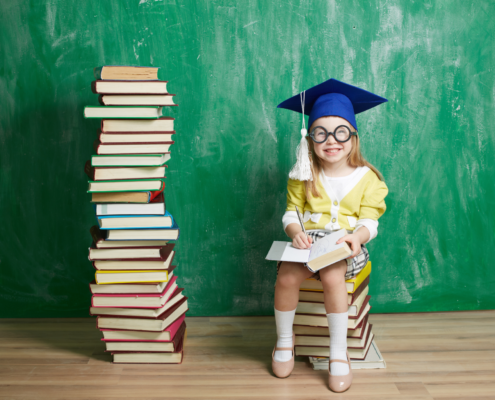 Young girl sits on stack of books next to a stack of books wearing a graduation cap