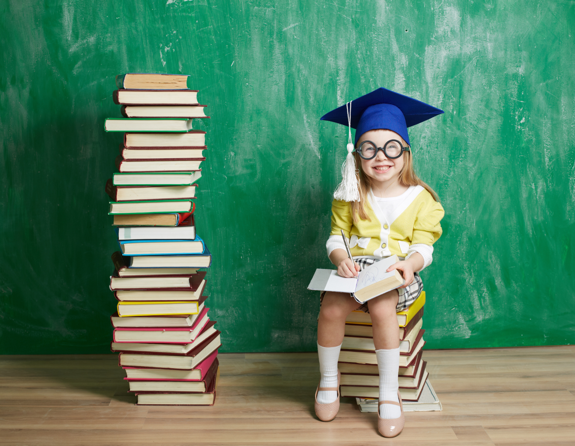 Young girl sits on stack of books next to a stack of books wearing a graduation cap 