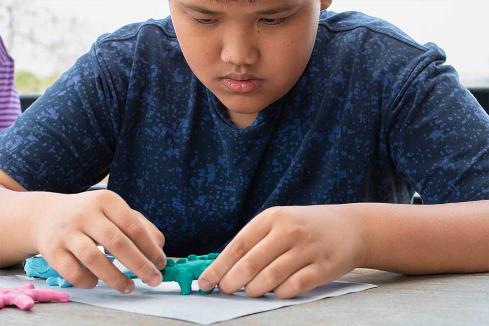 An autistic boy molds green model clay into the shape of a dinosaur.
